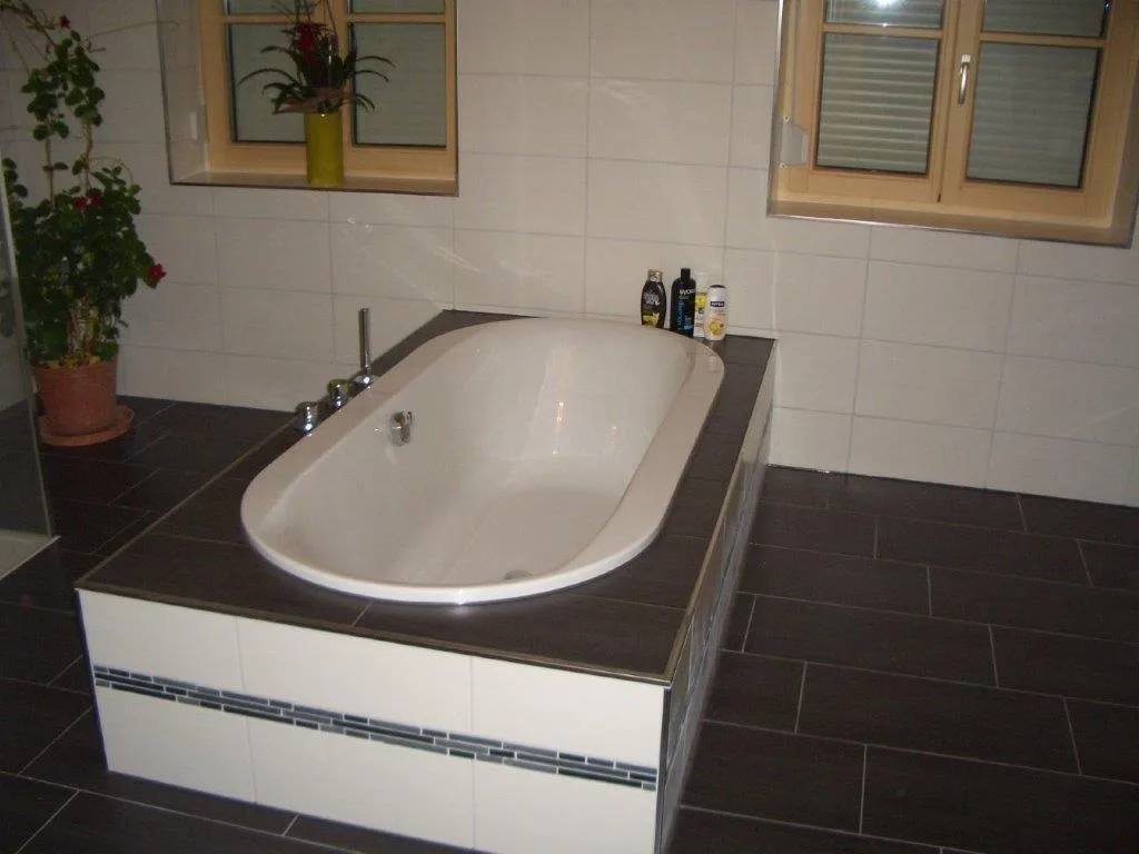 Eingelassene Badewanne in einer gefliesten Umrandung mit weißer Front und dunklem Bodenbelag